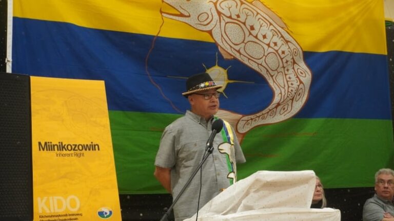 A man stands on a podium with a First Nation flag behind him.