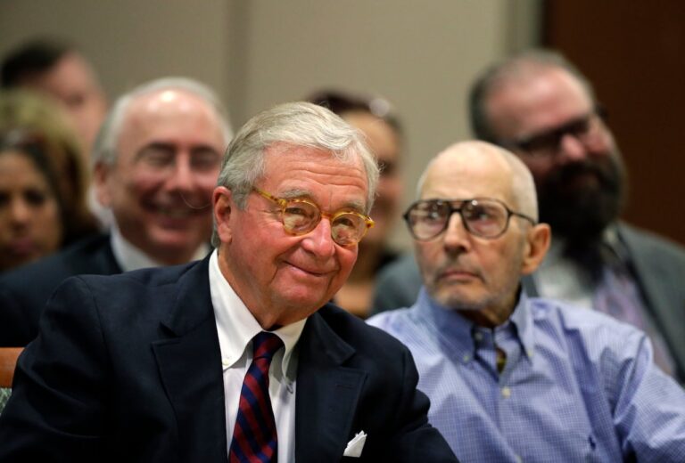 A gray-haired, clean-shaven man in a suit and tie smiles while sitting next to a pale, older man.